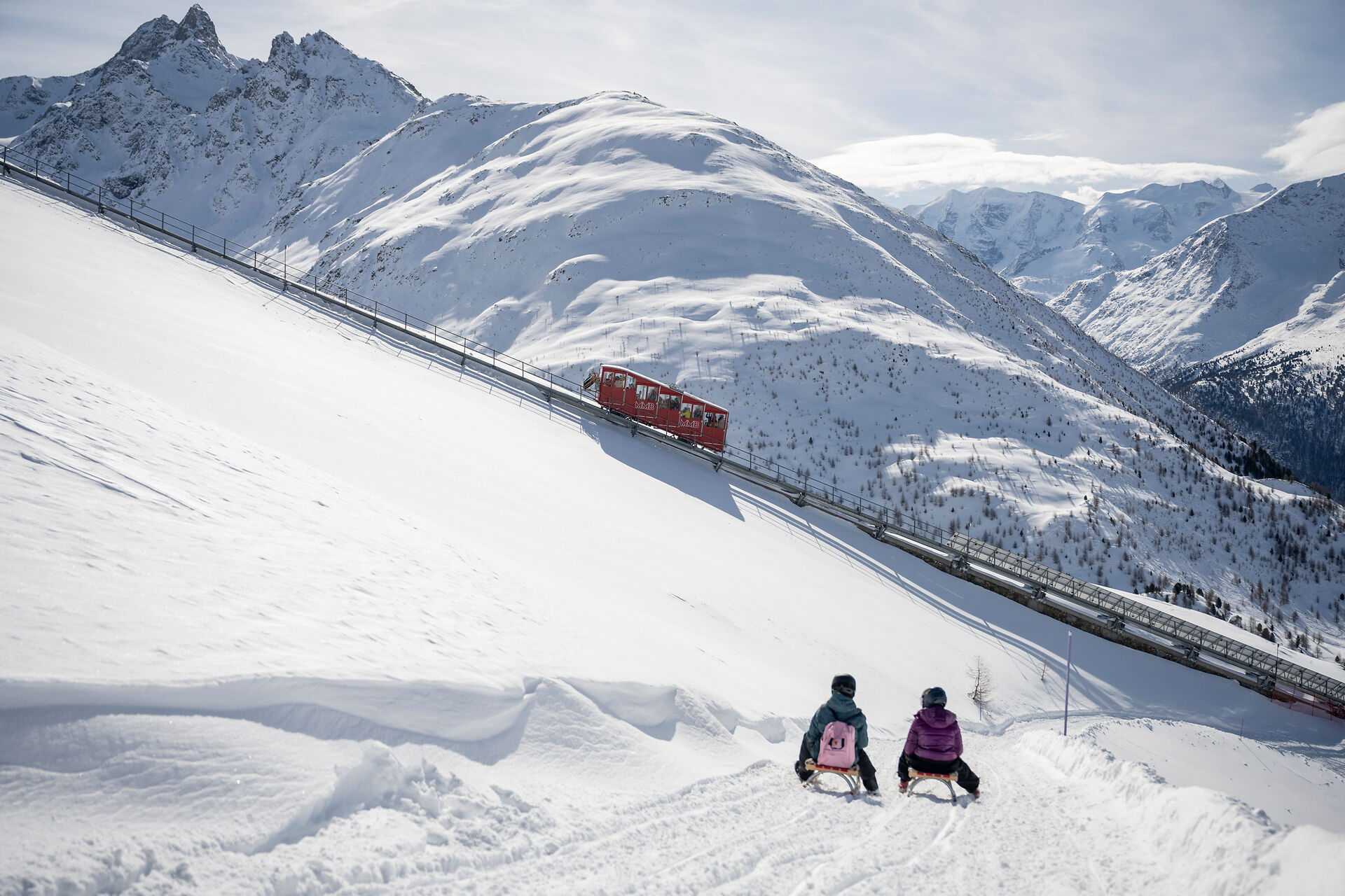 Zwei Personen mit Helmen und Winterbekleidung sitzen auf Holzschlitten und fahren einen verschneiten Hang hinunter. Vor ihnen erstreckt sich die weitläufige, verschneite Berglandschaft des Engadins mit der historischen, roten Standseilbahn Muottas Muragl, die durch das Bild fährt.