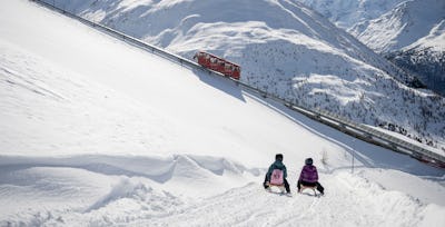 Schlitteln | Muottasmuragl Bergerlebnis | Winter Zwei Personen mit Helmen und Winterbekleidung sitzen auf Holzschlitten und fahren einen verschneiten Hang hinunter. Vor ihnen erstreckt sich die weitläufige, verschneite Berglandschaft des Engadins mit der historischen, roten Standseilbahn Muottas Muragl, die durch das Bild fährt.