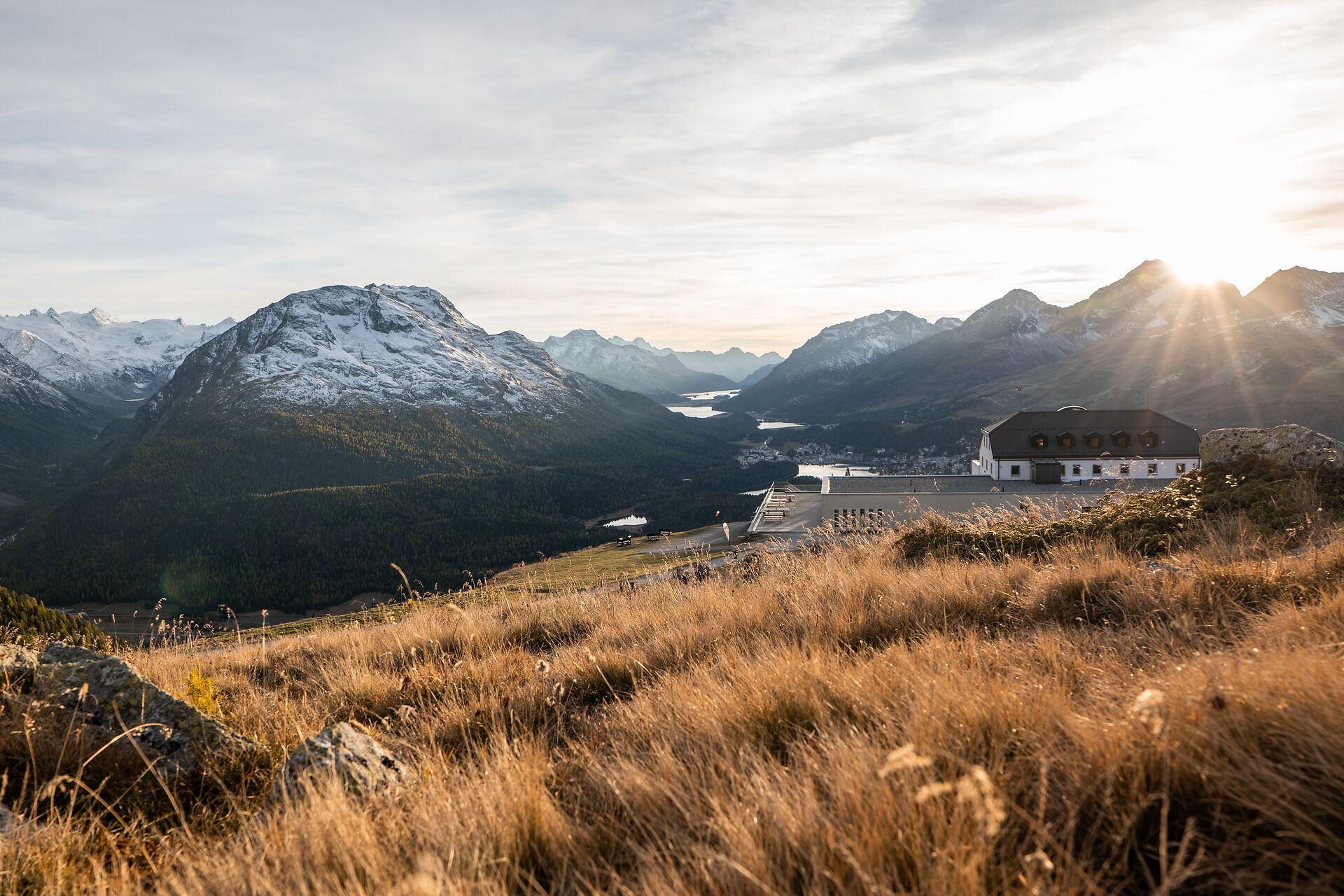 Herbstliche Berglandschaft auf Muottas Muragl mit goldenem Gras und Felsen im Vordergrund. Im Hintergrund sind verschneite Berggipfel, bewaldete Berghänge und mehrere Seen zu sehen. Rechts steht das Gebäude des Romantik Hotel Muottas Muragl, während die tief stehende Sonne über den Bergen durch die Wolken scheint.