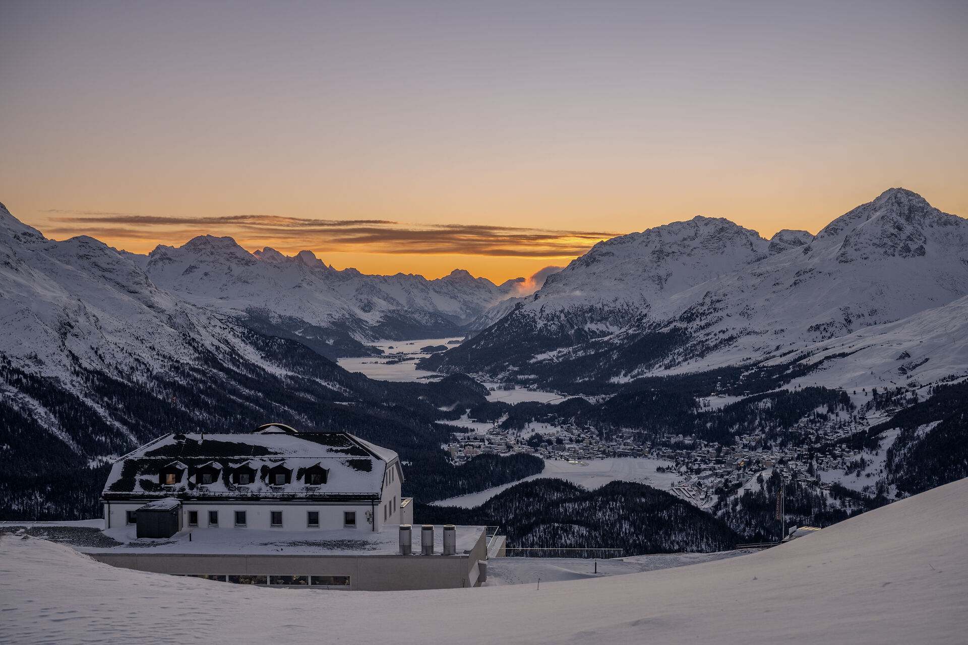 Talblick von oberhalb des Romantik Hotel Muottas Muragl auf das verschneite Oberengadin mit seinen Seen.dahinter erstrecken sich verschneite Berghänge, Wälder, zugefrorene Seen und eine weitläufige alpine verschneite Bergkulisse mit Gelb-Rotem Sonnenuntergang.