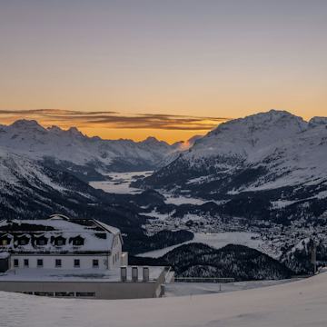 Romantik Hotel Muottas Muragl - Sonnenuntergang - Winter Talblick von oberhalb des Romantik Hotel Muottas Muragl auf das verschneite Oberengadin mit seinen Seen.dahinter erstrecken sich verschneite Berghänge, Wälder, zugefrorene Seen und eine weitläufige alpine verschneite Bergkulisse mit Gelb-Rotem Sonnenuntergang.