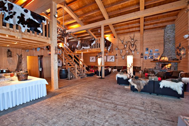 Rustikal eingerichteter Innenraum der Chesa Marguns mit Holzwänden, Holzbalken und einer Galerie mit Holztreppe. Mehrere Sitzgruppen mit dunklen Sofas und Sesseln sind mit Fellbezügen und Fellen dekoriert. An den Wänden hängen Geweihe, Fotografien und Bilder; rechts befindet sich ein Natursteinkamin mit brennendem Feuer. Links steht ein gedeckter Tisch mit Gläsern und Getränken.