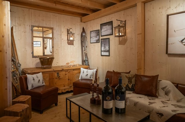 Rustikal eingerichtete Sitzecke der Chesa Marguns mit braunen Ledersesseln und einem Sofa mit gemusterten Kissen und Decke. Vor dem Sofa steht ein niedriger Tisch mit zwei Weinflaschen und Kerzenständern. Im Hintergrund sind eine Holzkommode, alte Skier, gerahmte Fotografien und weitere alpine Dekorationen zu sehen.