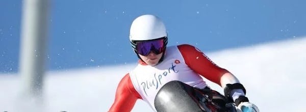 Para Ski - PluSport Skifahrer mit Monoskibob in roter Skibekleidung und weissem Helm fährt auf einer verschneiten Skipiste auf der Corviglia durch ein blau markiertes Tor.