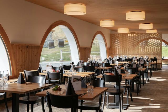 Das Restaurant Marguns ist für die Metzgeta mit mehreren gedeckten Holztischen, schwarzen Stühlen und gefalteten schwarzen Servietten hergerichtet. Grosse Rundbogenfenster und eine dekorative Holzverkleidung prägen den hellen Innenraum.