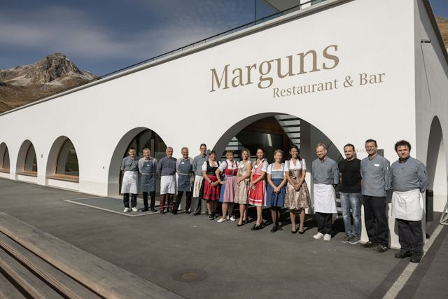 Gruppenfoto des Teams vor dem Restaurant & Bar Marguns. Mehrere Mitarbeitende stehen in Arbeitskleidung sowie teilweise in Tracht vor dem Gebäude mit grossen Rundbogenfenstern. Im Hintergrund ist ein Berggipfel des Corviglia-Gebiets zu sehen.