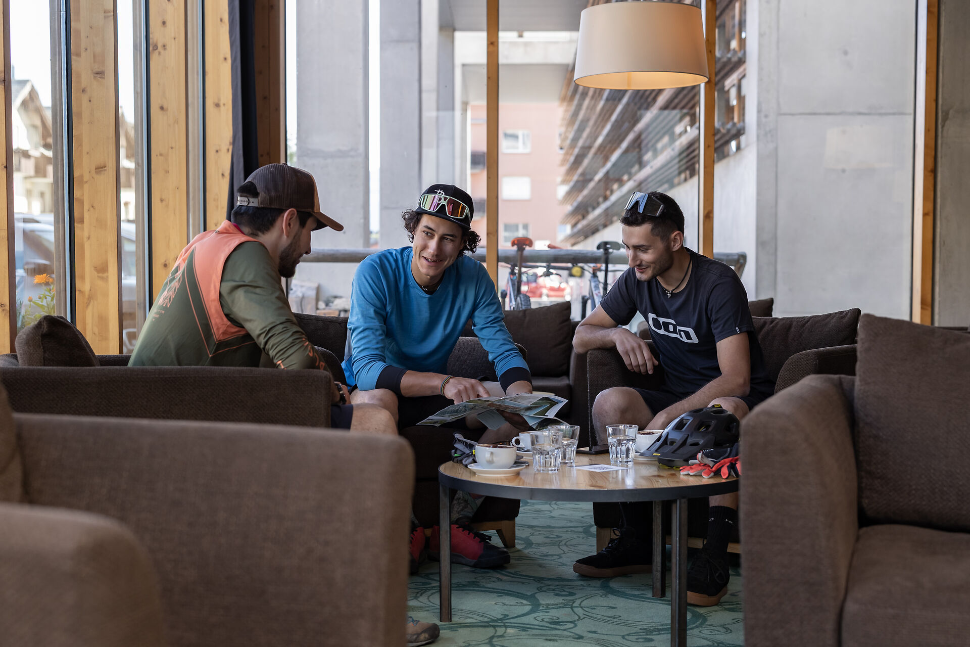 Vor einer Mountainbiketour auf Corviglia sitzen drei Männer im modern eingerichteten Innenbereich des ALL IN ONE HOTEL INN LODGE an einem runden Holztisch. Bei Getränken planen sie ihre Tour mithilfe einer Karte.