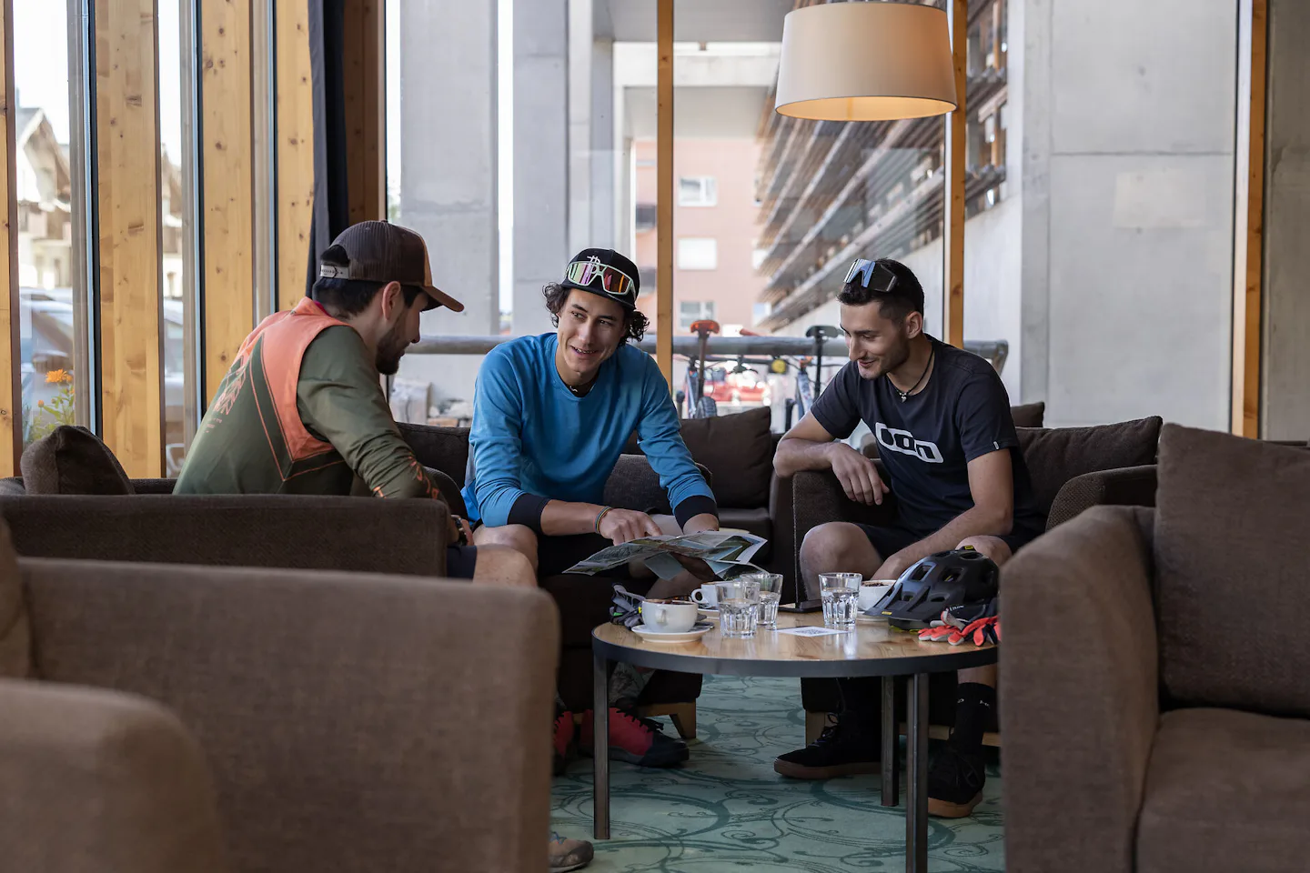 Mountainbiker - ALL IN ONE HOTEL INN LODGE Vor einer Mountainbiketour auf Corviglia sitzen drei Männer im modern eingerichteten Innenbereich des ALL IN ONE HOTEL INN LODGE an einem runden Holztisch. Bei Getränken planen sie ihre Tour mithilfe einer Karte.