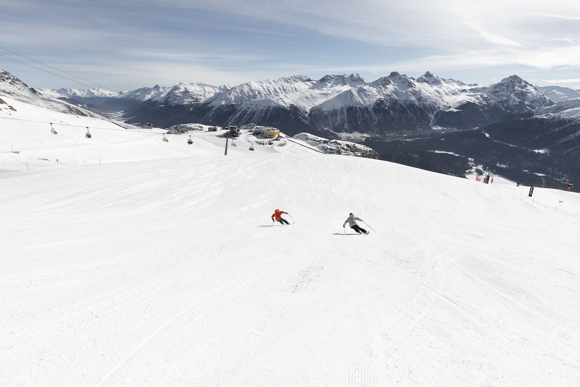 Auf einer breiten, präparierten Skipiste fahren zwei Skifahrer den Hang Richtung der gelben Bergstation Corviglia hinunter. Im Hintergrund sind eine Sesselbahn, die Bergstationen Corviglia und die weitläufige verschneite alpine Berglandschaft des Engadins mit bewaldeten Hängen und Berggipfeln zu sehen.
