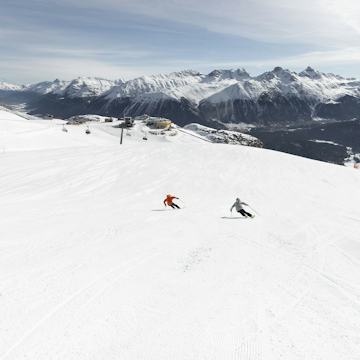 Skifahren Corviglia Auf einer breiten, präparierten Skipiste fahren zwei Skifahrer den Hang Richtung der gelben Bergstation Corviglia hinunter. Im Hintergrund sind eine Sesselbahn, die Bergstationen Corviglia und die weitläufige verschneite alpine Berglandschaft des Engadins mit bewaldeten Hängen und Berggipfeln zu sehen.