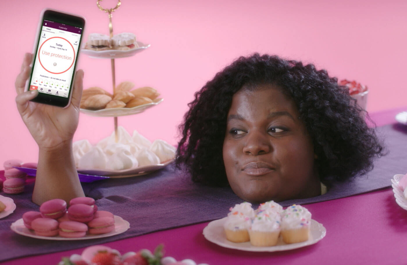 Woman with her head sticking through a table laden with cake holding a phone which has a screen showing the text 'not fertile'.