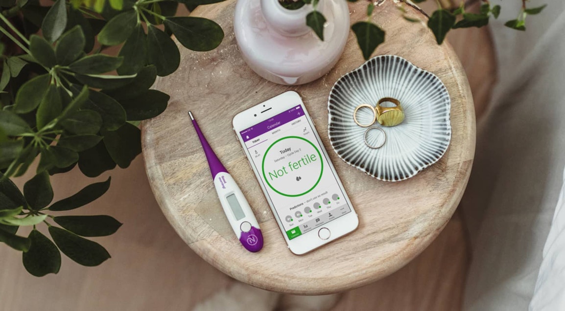 The Natural Cycles app and thermometer on a wooden table next to a vase and a dish containing some jewellery.