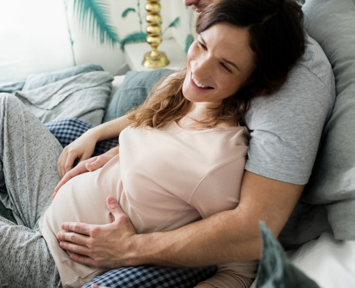woman leaning on her partner in bed while he is holding her pregnant belly
