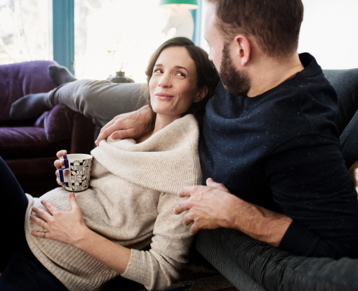 couple lying in their sofa while the woman is drinking tea and holding her pregnant belly