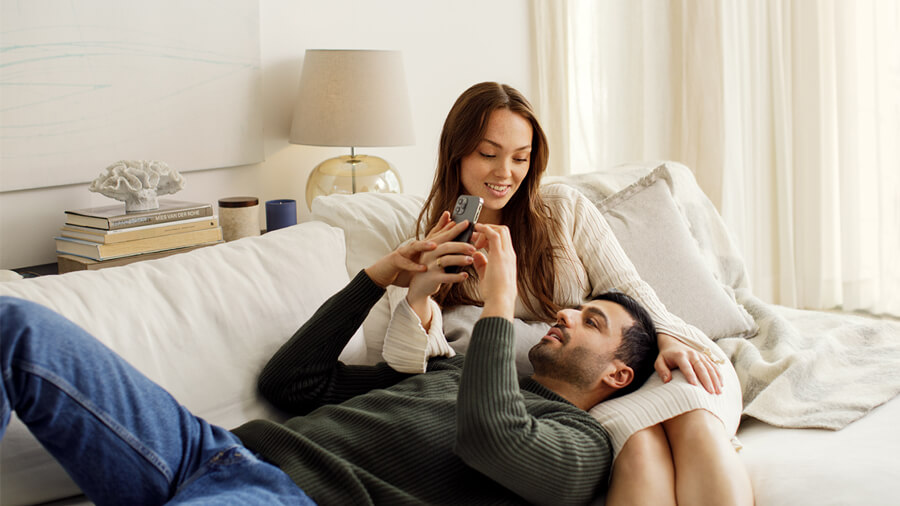 Man lying on the sofa with his head in his partern's lap, they're both looking at a phone