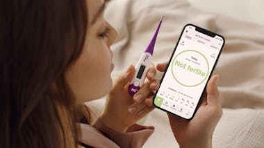 Woman holding thermometer and looking at phone screen that says 'not fertile'