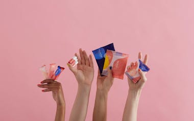 Four hands holding Callaly's period products against an empty pink background. From left to right: tampliners, tampons, pads and liners.