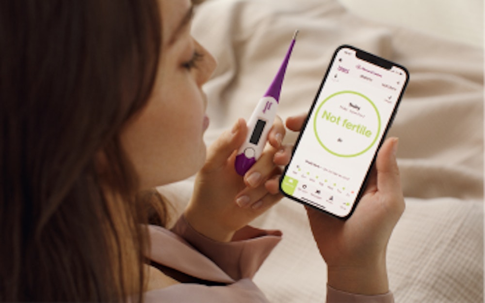 Woman holding a thermometer and smartphone displaying the Natural Cycles app