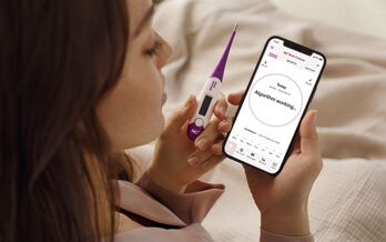 Woman holding a thermometer and smartphone displaying the Natural Cycles app