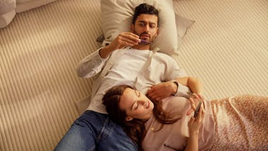 Man and woman lying on the carpet, he's looking at a thermometer, she's looking at her phone.