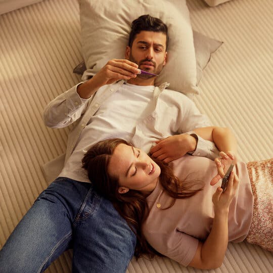 Casal deitado com travesseiros, um termômetro e um smartphone