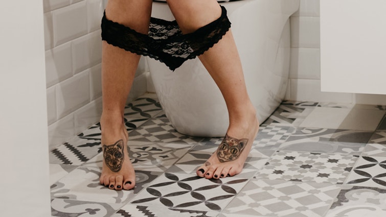 Legs of a woman sat on the toilet with black lace underwear around her ankles and two cat tattoos on her feet