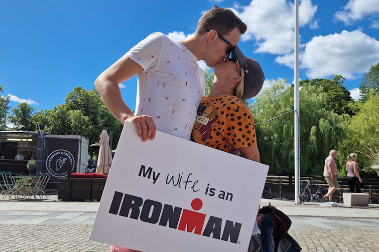 Manu kissing her husband who's holding a sign that says 'my wife is an Ironman' in a sunny square