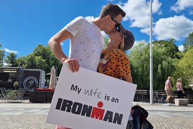 Manu kissing her husband who's holding a sign that says 'my wife is an Ironman' in a sunny square