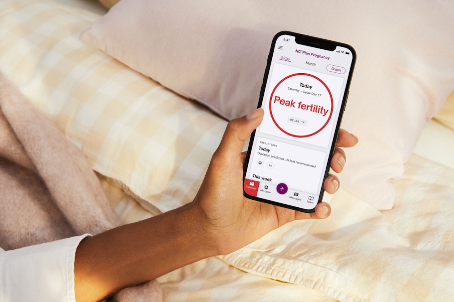 Hand holding phone on yellow bedsheets the phone screen has a red circle with the text 'peak fertility' in the center