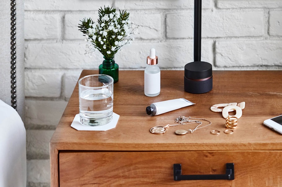 Bedside table showing a glass of water, a tube of cream, rings, necklaces, flowers and a phone