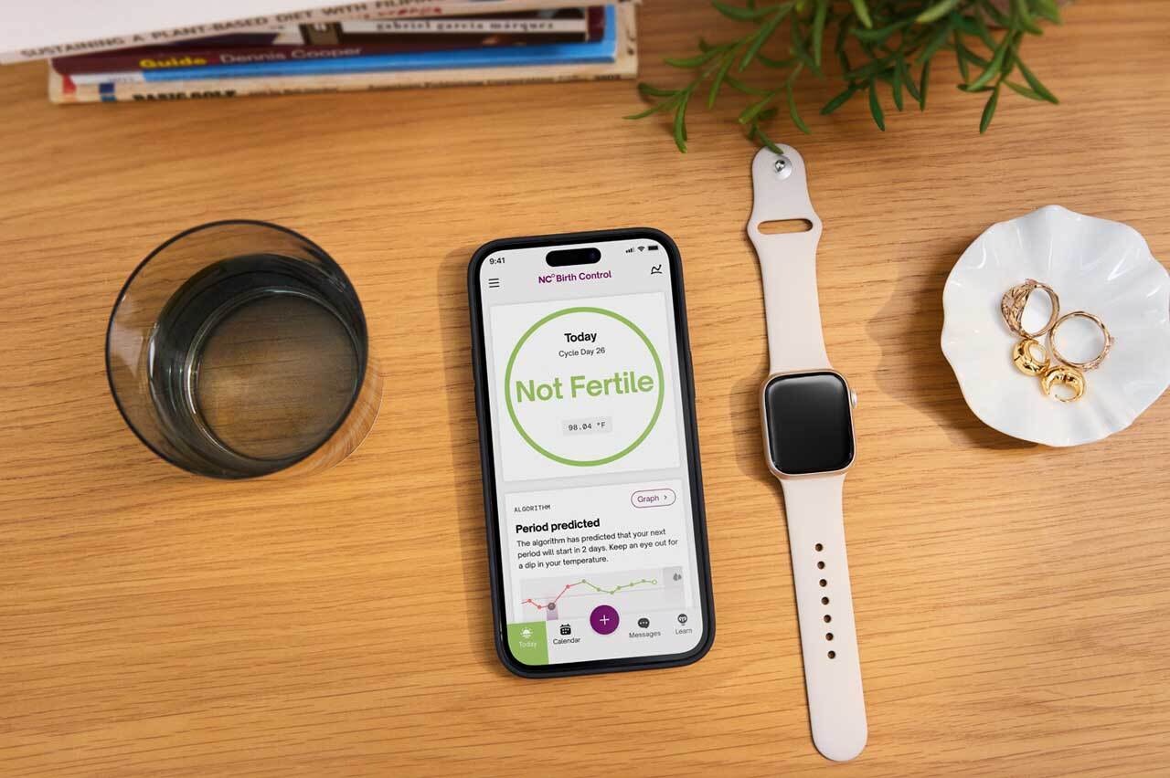 Photograph of Apple Watch next to phone showing Natural Cycles app screen: not fertile. Both are sat on a wooden table with books, a plant, a glass of water and some rings.