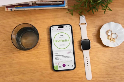 Photograph of Apple Watch next to phone showing Natural Cycles app screen: not fertile. Both are sat on a wooden table with books, a plant, a glass of water and some rings.