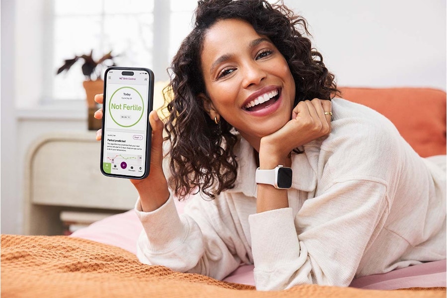 Kvinna ligger pÄ mage pÄ en sÀng med en Apple Watch pÄ armen och hÄller upp en telefon med Natural Cycles-appen öppen för kameran