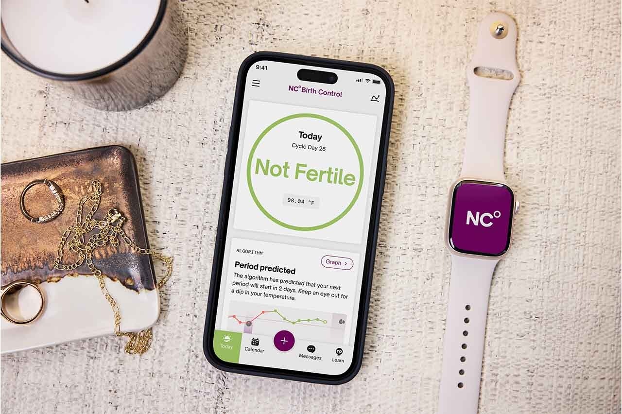 Apple Watch on a table displaying the NC logo next to a phone showing the text 'not fertile'