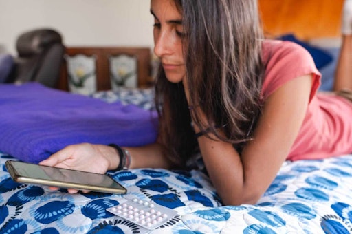 Woman lying on bed looking at her phone with a packet of birth control pills beside her