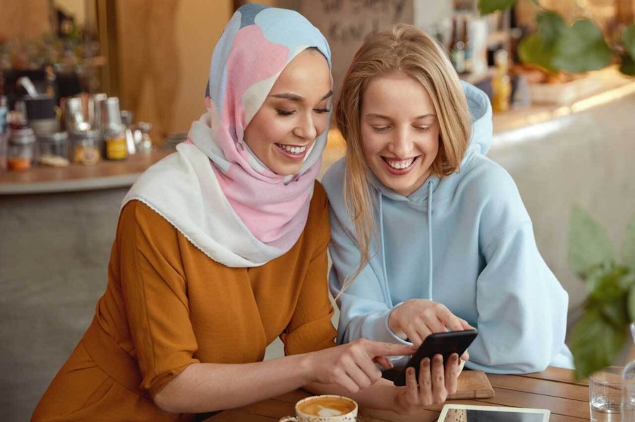 Two women looking at a phone, smiling, in the kitchen with coffee