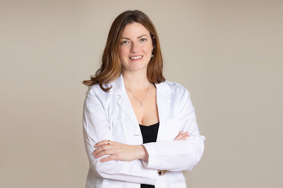 Dr Kerry Krauss stands with her arms folded smiling at the camera. She is wearing a white doctor's coat over a black top, the background is a neutral beige..