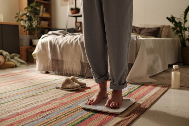 Woman in her bedroom, shown from the waist down standing on scales wearing sweatpants