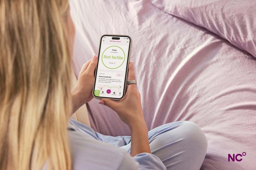 Woman wearing an Oura Ring sitting on lilac bedsheets looking at her phone which displays the text 'Not Fertile' from the Natural Cycles app