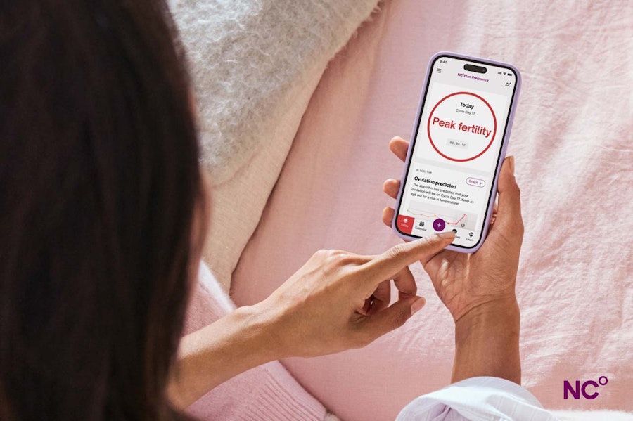 Woman on pink bedsheets holding a phone with the fertility status 'peak fertility' as shown in the Natural Cycles app