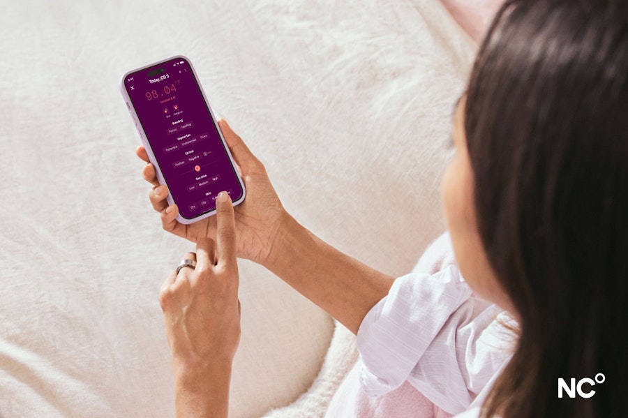 Woman sat on cream bedsheets, holding her phone and tracking cycle symptoms in the Natural Cycles app