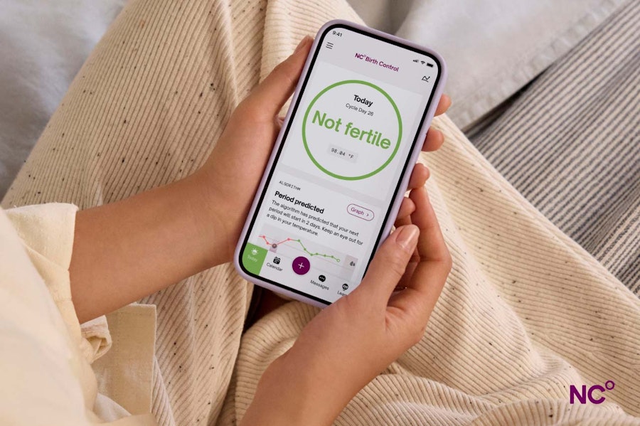 Person sitting on bed holding a phone which shows the Natural Cycles app with text 'Not fertile'