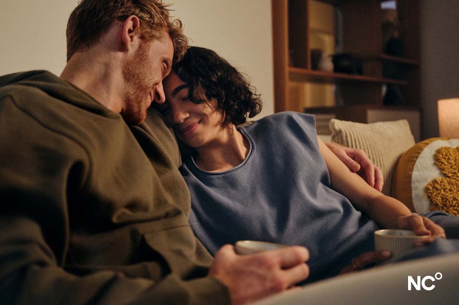 Couple sat on the sofa holding cups of coffee and cuddling