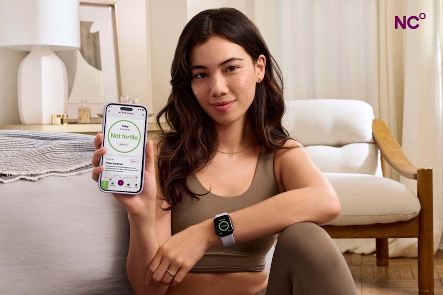 Woman in yoga gear sat on the floor, holding her phone which shows the Natural Cycles app and the text 'not fertile'