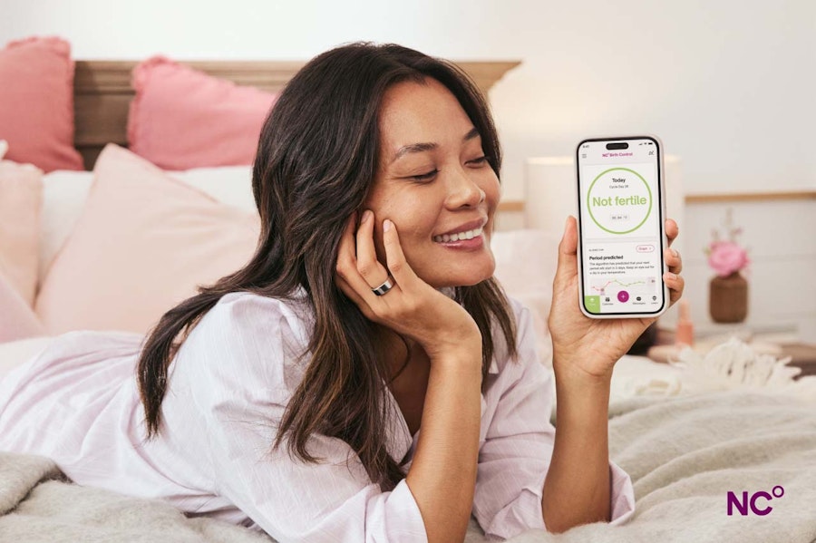 Woman lying on her bed smiling, wearing Oura Ring and holding her phone which shows the Natural Cycles app and the text 'Not Fertile'