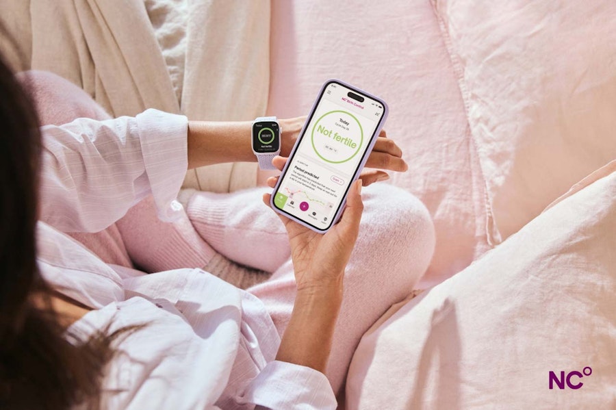 Woman sat on pink bedding, wearing an Apple Watch and holding her phone with the Natural Cycles app open and the text 'Not Fertile' displayed on the screen