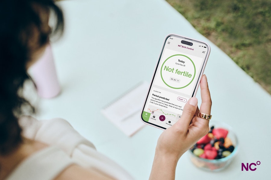 Woman wearing an Oura Ring while standing outside and holding her phone above a table with a bowl of fresh fruit on it. The phone is open on the Natural Cycles app and shows the text 'Not fertile'