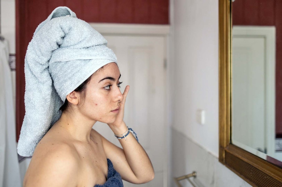 Woman with her hair tied up with a towel standing in front of her bathroom mirror looking into it while touching her face
