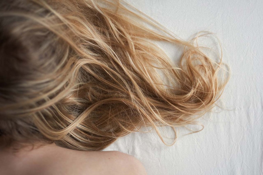 Woman lying on her bed face down, her face is not visible, you can only see her blonde hair falling onto the white sheets
