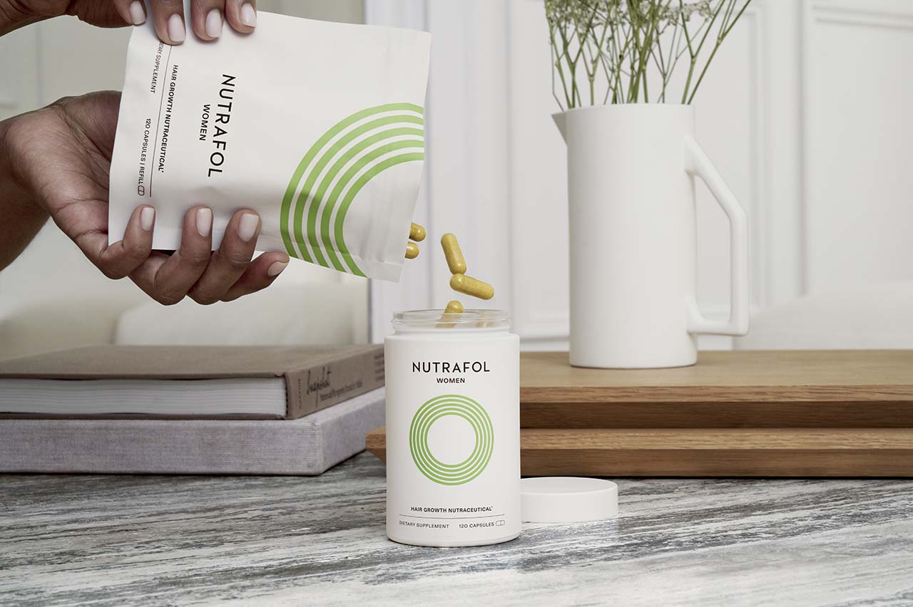 Photograph of hands refilling a container branded Nutrafol Woman, the container sits on a wooden surface with a white vase of flowers in the background. 